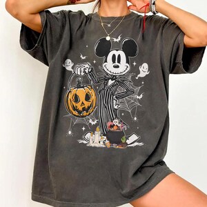 May include: Dark gray t-shirt featuring a Halloween-themed design. The graphic depicts Mickey Mouse dressed as Jack Skellington, holding a carved pumpkin. The design includes ghosts, bats, and spiderwebs, creating a spooky aesthetic.