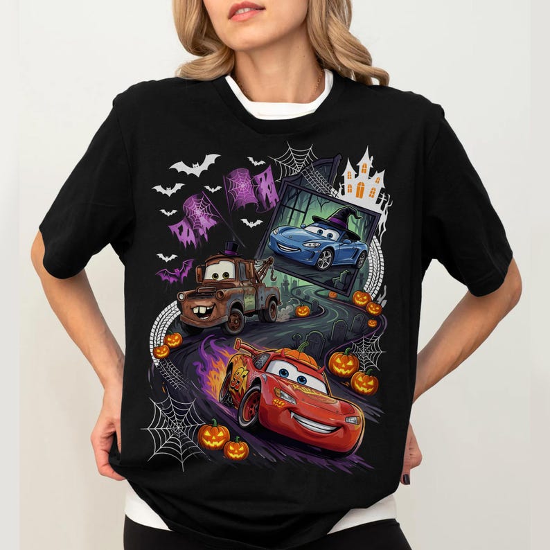K&ouml;nnte beinhalten: Ein schwarzes Kurzarm-T-Shirt mit einem Grafikdruck von Charakteren aus dem Film Cars in einer Halloween-Umgebung. Das Design zeigt einen braunen Abschleppwagen, ein rotes Rennauto, ein blaues Auto mit Hexenhut, Flederm&auml;use, Spinnennetze und K&uuml;rbisse.