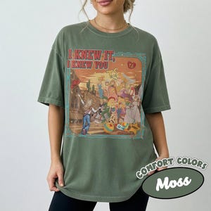 Op de afbeelding: Een mosgroen t-shirt met korte mouwen en een grafische print van een groep stripfiguren in een woestijnomgeving. De tekst "I KNEW IT, I KNEW YOU" staat in het rood boven de illustratie, die een gitaar en een hartpictogram bevat.