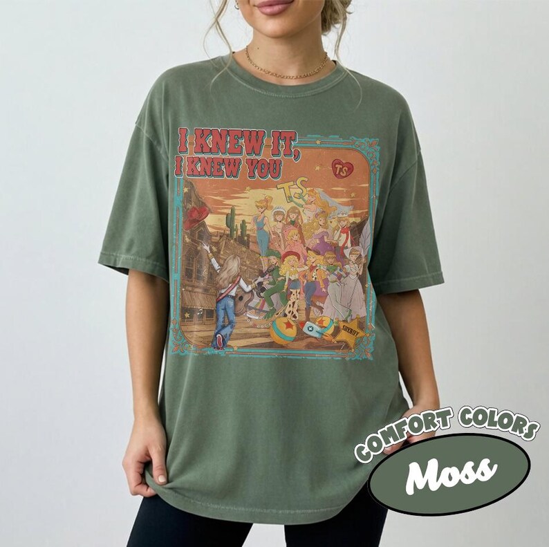 Op de afbeelding: Een mosgroen t-shirt met korte mouwen en een grafische print van een groep stripfiguren in een woestijnomgeving. De tekst "I KNEW IT, I KNEW YOU" staat in het rood boven de illustratie, die een gitaar en een hartpictogram bevat.