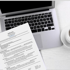 Puede incluir: Un portátil, un currículum vitae y una taza de café sobre una superficie de madera blanca. El currículum vitae se titula "Emily Green" y enumera la experiencia profesional. El portátil está abierto y la taza de café es blanca.