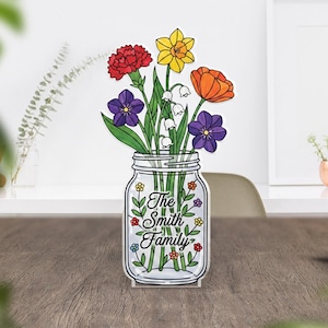 Könnte beinhalten: Eine transparente Acrylvase in Form eines Einmachglases, gefüllt mit bunten Blumen. Die Blumen umfassen rote Nelken, gelbe Narzissen, orangefarbene Tulpen, violette Veilchen und weißes Maiglöckchen. Der Text "The Smith Family" ist auf dem Glas abgebildet.