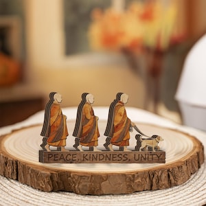 May include: Wooden decorative piece featuring three figures in robes, one walking a dog. The figures are layered with various wood tones. The base reads "PEACE, KINDNESS, UNITY". The piece is displayed on a wooden slice.