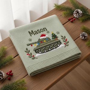 May include: A sage green knitted blanket featuring an embroidered design of a military tank wearing a Santa hat and decorated with colorful Christmas lights. The name Mason is embroidered above the tank, accompanied by a small Christmas tree.