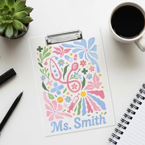 May include: White clipboard with a colorful floral design featuring a stethoscope, band-aid, and syringe. The name "Ms. Smith" is printed in blue. A cup of coffee, a succulent plant, a pen, and a notebook are also visible.