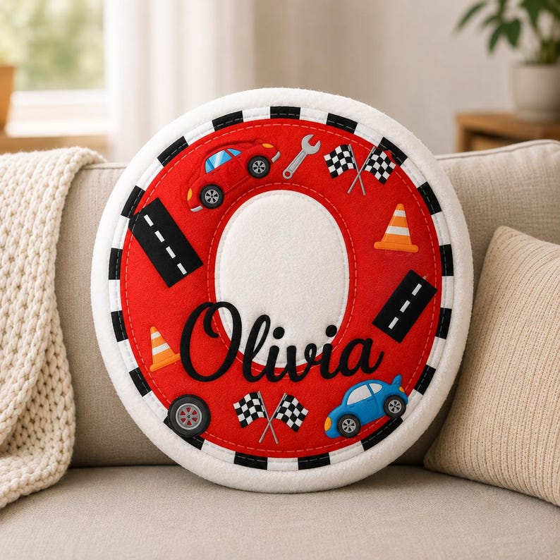 May include: A round, red and white felt pillow featuring a racing theme with appliqu&eacute;s of a red car, a blue car, checkered flags, traffic cones, a wrench, and road segments. The name Olivia is embroidered in black script across the center.