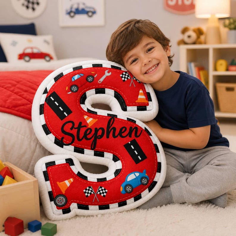 May include: A plush letter S pillow in bright red featuring a black and white checkered border. The surface is decorated with printed graphics of cars, traffic cones, a wrench, and checkered flags. The name Stephen is written in black script.