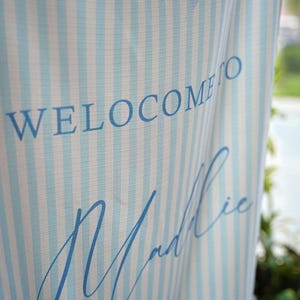 K&ouml;nnte beinhalten: Ein vertikales Stoffbanner mit einem hellblau-wei&szlig;en vertikalen Streifenmuster. Der in Blau gedruckte Text lautet WELCOME TO, gefolgt von dem Namen Maddie in Schreibschrift und dem Wort AND darunter in einer Blockschrift.