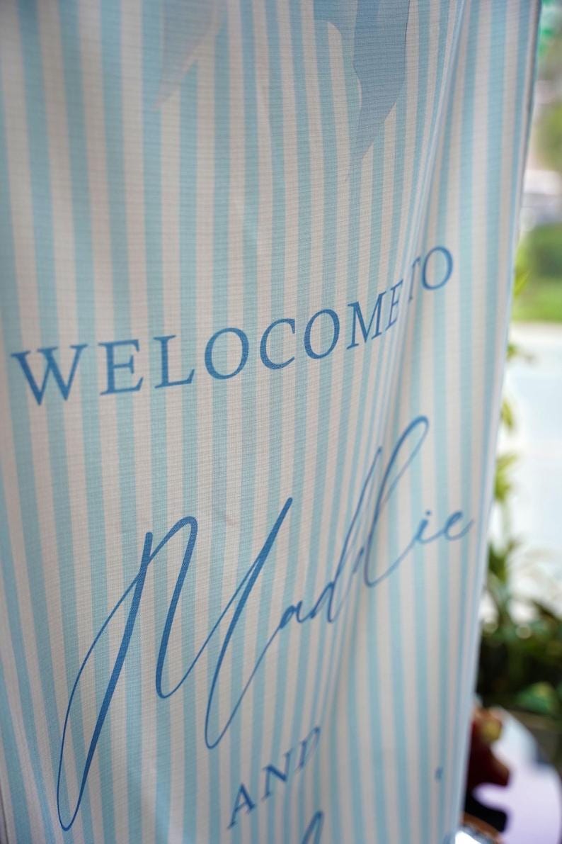K&ouml;nnte beinhalten: Ein vertikales Stoffbanner mit einem hellblau-wei&szlig;en vertikalen Streifenmuster. Der in Blau gedruckte Text lautet WELCOME TO, gefolgt von dem Namen Maddie in Schreibschrift und dem Wort AND darunter in einer Blockschrift.