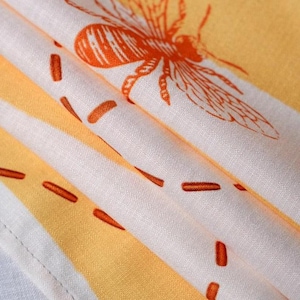K&ouml;nnte beinhalten: Ein gefaltetes Textil mit einem sich wiederholenden Muster aus orangefarbenen Bienen und gestrichelten Linien auf einem wei&szlig; und hellgelb gestreiften Hintergrund. Der Stoff zeigt eine sichtbare Webstruktur mit einem Saum am unteren Rand.