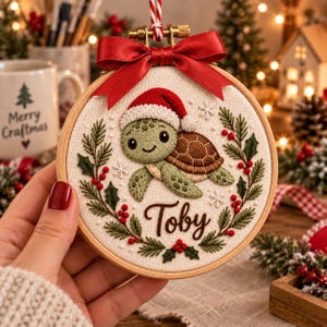 Könnte beinhalten: Ein runder Holzstickrahmen mit einem Stoffeinsatz, der eine gestickte grüne Schildkröte mit roter Weihnachtsmütze zeigt. Die Schildkröte ist von einem Kranz aus grünen Tannenzweigen und roten Beeren umgeben, mit dem Namen Toby in brauner Schrift.