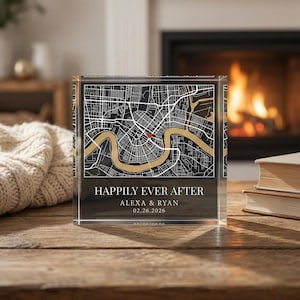 Puede incluir: Un bloque acrílico transparente con un diseño de mapa en negro, dorado y blanco. El mapa tiene un marcador rojo. Se muestra el texto "HAPPILY EVER AFTER", junto con nombres y una fecha. El bloque está sobre una superficie de madera.