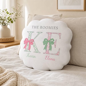 Op de afbeelding: Een wit kussen met geschulpte randen en de tekst THE ROOMIES bovenaan. Daaronder staan de grote letters K en E met bloemmotieven. De K is groen met een roze strik en de E is roze met een groene strik. De namen Kevin en Elena staan eronder gedrukt.