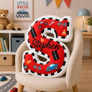 May include: A plush letter S pillow in bright red with a white and black checkered border. The surface features printed graphics of blue and red race cars, orange traffic cones, black road segments, wrenches, and checkered flags with the name Stephen.