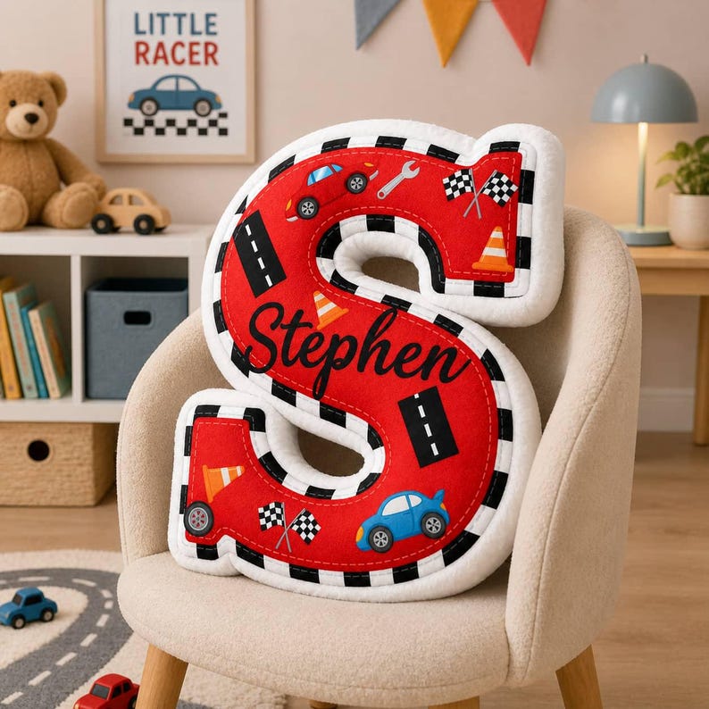 May include: A plush letter S pillow in bright red with a white and black checkered border. The surface features printed graphics of blue and red race cars, orange traffic cones, black road segments, wrenches, and checkered flags with the name Stephen.