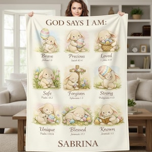 May include: A cream-coloured blanket with illustrations of bunnies holding Easter eggs and flowers. The text "GOD SAYS I AM: Brave, Precious, Loved, Safe, Forgiven, Strong, Unique, Blessed, Known" is displayed, along with the name "SABRINA".