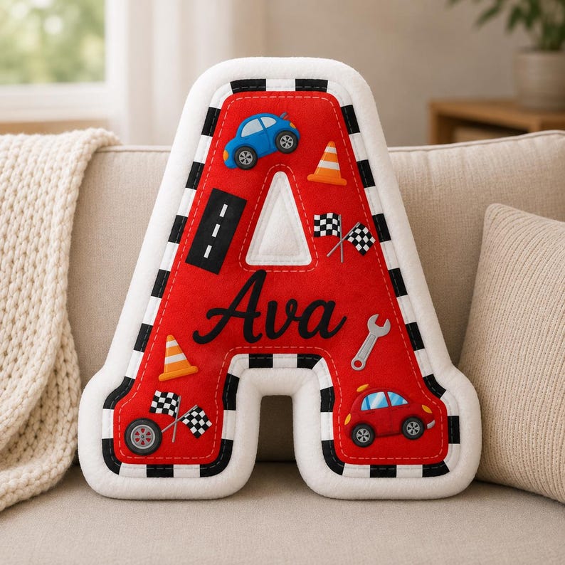May include: A red letter A-shaped pillow featuring a black and white checkered border. The pillow is decorated with appliqu&eacute;s of two cars, two traffic cones, two sets of checkered flags, a road strip, and a wrench. The name Ava is embroidered in black.