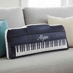 May include: A decorative pillow shaped like a black and white keyboard, with the name "Alyssa" in cursive script. The pillow is rectangular and rests on a gray sofa, with other pillows in the background.