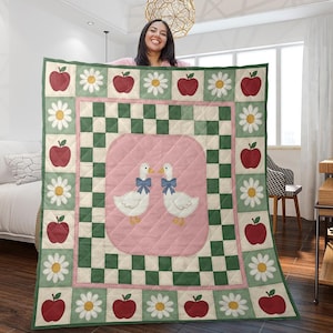 Op de afbeelding: Een gewatteerde deken met een roze middenstuk met twee ganzen met blauwe strikken. De rand heeft afwisselend rode appels en witte madeliefjes, met een groen en wit geblokt patroon. De deken wordt door een persoon omhoog gehouden.