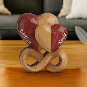 May include: A wooden sculpture featuring three heart-shaped elements, two in burgundy and one in natural wood, atop an infinity symbol base. The burgundy hearts are inscribed with names, and the base displays a date. A decorative item.