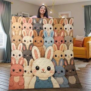 Puede incluir: Una manta acolchada con un patrón repetido de conejitos de dibujos animados en varios colores, incluyendo beige, rosa, gris y azul. Los conejitos tienen ojos grandes y llevan monos. La manta se muestra en interiores, con una persona sosteniéndola.