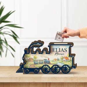 Peut inclure: Une tirelire en bois en forme de train, de couleur bleue et marron. Elle présente une peinture à l'aquarelle d'un train et d'un paysage. Le nom "ELIAS Monroe" est imprimé sur la tirelire.