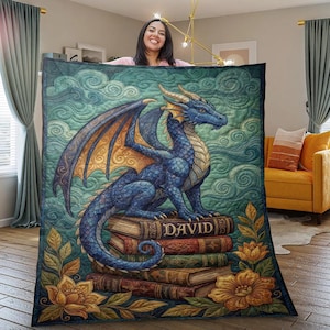 Puede incluir: Una manta de dragón azul con una ilustración detallada. El dragón está posado sobre una pila de libros con el nombre "DAVID" en el libro superior. La manta tiene un fondo verde azulado con detalles florales.