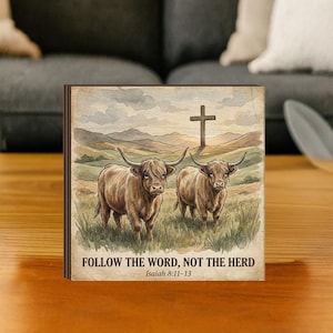 May include: A square wooden block featuring a watercolor-style illustration of two Highland cows in a grassy field with a cross in the background. The text "FOLLOW THE WORD, NOT THE HERD" and Isaiah 8:11-13 are printed at the bottom.