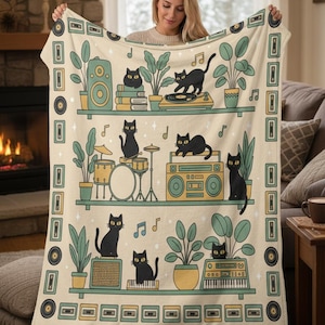 May include: A cream-colored throw blanket featuring a whimsical design of black cats on shelves with musical instruments, plants, and cassette tapes. The blanket has a decorative border with musical notes and cassette tape icons.