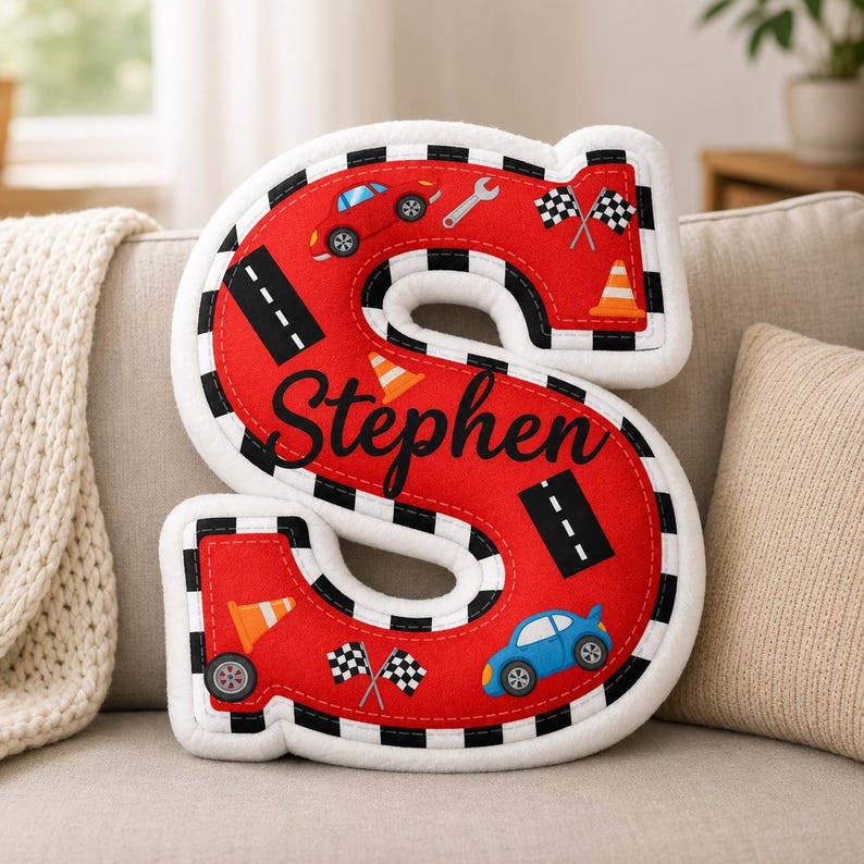 May include: A plush letter S pillow in red with a black and white checkered border. The surface features a road pattern, orange traffic cones, checkered flags, a wrench, and two small cars. The name Stephen is embroidered in black script.