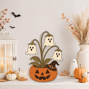 Puede incluir: Un soporte decorativo de acrílico con una ilustración central de una maceta de calabaza naranja tallada con un sombrero de bruja negro, que contiene una planta con tres flores blancas en forma de fantasma con ojos oscuros y bocas abiertas.