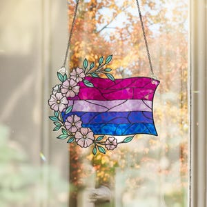 Peut inclure: Un drapeau de la fierté bisexuelle en vitrail, avec des rayures roses, violettes et bleues, orné de fleurs roses et de feuilles vertes. Le drapeau est suspendu par une chaîne argentée, créant une décoration de fenêtre.