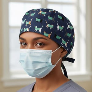 May include: A navy blue surgical scrub cap featuring a repeating pattern of stylized cats in various colors. The cats are depicted in green, orange, white, and black. The cap is tied at the back. A light blue surgical mask is worn.