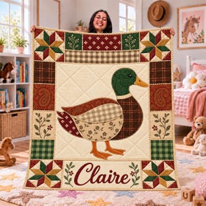 May include: A quilted blanket featuring a central mallard duck appliqué on a cream background, surrounded by a patchwork border of stars, plaid, and floral patterns in red, green, and brown. The name Claire is embroidered at the bottom.