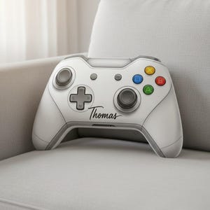 May include: A white and gray video game controller with the name "Thomas" in script. The controller has colored buttons and directional controls. It rests on a gray sofa.