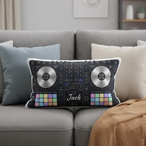 May include: A rectangular pillow featuring a DJ mixer design with two silver-colored turntables and colorful buttons. The name "Jack" is printed in white. The pillow is on a gray couch with other pillows.