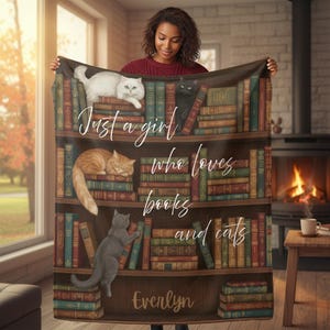 Może przedstawiać: Przytulny koc z motywem półki na książki, z kotami i książkami. Koc ma napis "Just a girl who loves books and cats" i spersonalizowane imię. Koty są białe, pomarańczowe i szare.