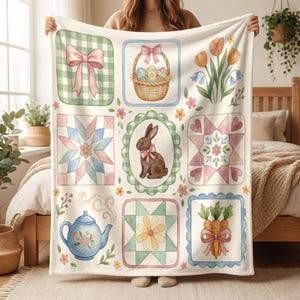 May include: A cream-coloured blanket with a patchwork design featuring Easter-themed illustrations. The squares include a gingham bow, a basket of eggs, tulips, a bunny, and a teapot. The blanket is held up by a person.