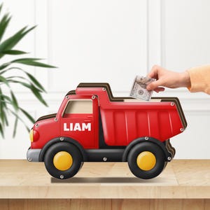 May include: A red toy dump truck money bank with the name "LIAM" in white letters on the side. The truck has black wheels with yellow centres. A hand is placing a US $100 bill into the slot on top of the truck.