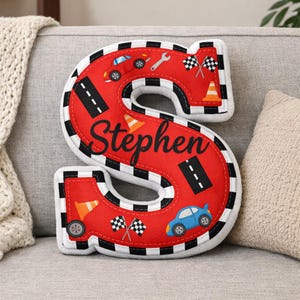 Può includere: Un cuscino a forma di lettera "S" con un tema di auto da corsa. Il cuscino rosso presenta bordi a scacchi bianchi e neri, grafica di auto da corsa e il nome "Stephen" in corsivo nero. Include bandiere da corsa, coni e illustrazioni di auto.