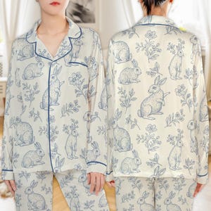 May include: White pajama set with a blue rabbit and floral pattern. The long-sleeved top has a notched collar and blue trim. The matching pants complete the sleepwear set. The fabric appears to be soft and silky.