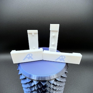 Könnte beinhalten: Eine 3D-gedruckte Skulptur mit einer blau-grauen geometrischen Basis, gekrönt von weißen rechteckigen Teilen. Die weißen Teile haben ein blaues "AR"-Logo. Die Skulptur steht vor einem dunklen Hintergrund.