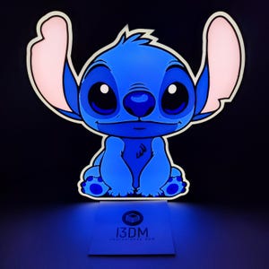 Op de afbeelding: Een blauwe cartoonkarakterlamp met grote oren en grote ogen. Het personage zit op een voet met de tekst "13DM" en het websiteadres. De lamp heeft een witte omtrek en roze accenten op de oren.