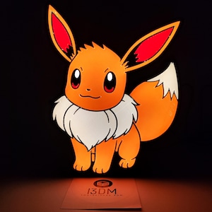 Puede incluir: Un personaje Eevee naranja de la franquicia Pokémon, con orejas con puntas rojas, un collarín blanco y una cola esponjosa. El personaje está sobre un fondo negro. Una tarjeta blanca con el texto "13DM Impresiones 3DM" está en la parte inferior.