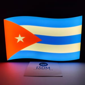 Puede incluir: Una bandera cubana iluminada, con un triángulo rojo y una estrella blanca, y rayas azules y blancas. La bandera está iluminada sobre un fondo negro. Una tarjeta blanca con el texto "I3DM Impresiones 3DM" está en la parte inferior.
