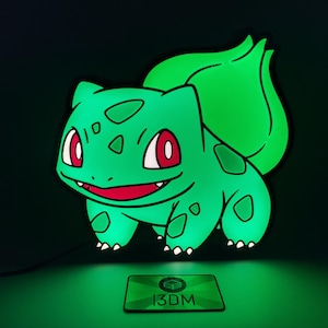 Könnte beinhalten: Ein grünes LED-Licht in Bulbasaur-Form mit roten Augen und weißen Zähnen. Das Licht hat eine schwarze Umrandung und befindet sich auf einem dunklen Hintergrund. Ein kleines grünes rechteckiges Schild mit dem Text "13DM" befindet sich unten.