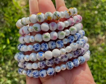 Pode incluir: Uma pilha de pulseiras com contas de cerâmica branca com padrões florais em amarelo, rosa e azul, intercaladas com contas de pérolas brancas. As pulseiras são seguradas em uma mão contra um fundo verde desfocado.