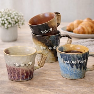 Puede incluir: Un conjunto de tazas de cerámica con superficies texturizadas y asas trenzadas. Las tazas presentan las palabras "Mummy", "Papa" y "Best Dad Ever". Las tazas son de color marrón, azul y beige, con un diseño rústico.