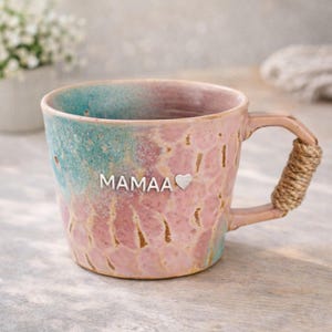 Könnte beinhalten: Eine Keramiktasse mit rosa und türkis Glasur. Die Tasse hat das Wort "Mamaa" und ein herzförmiges Detail. Der Griff ist mit Bindfaden umwickelt. Ein einzigartiger und dekorativer Artikel.