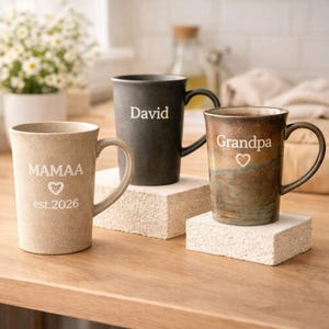 Könnte beinhalten: Drei Keramikbecher mit Henkeln werden auf einer Holzoberfläche präsentiert. Ein Becher ist beige mit "MAMAA" und "est.2026" aufgedruckt. Ein weiterer ist schwarz mit "David" aufgedruckt. Der dritte Becher ist braun mit "Grandpa" aufgedruckt.